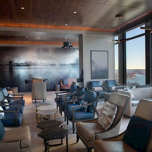 A modern lounge with comfortable seating, large windows offering a scenic view of the waterfront and Ferris wheel, and a mural on the back wall.