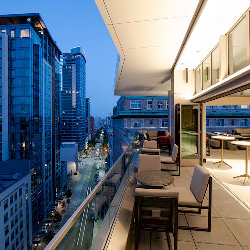 A modern balcony with seating overlooks a cityscape with tall buildings and illuminated streets, featuring a mix of natural and artificial light.