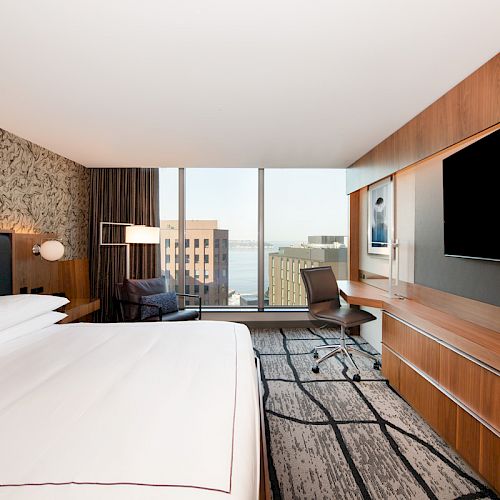 A modern hotel room with a large bed, wall-mounted TV, desk with chair, and window overlooking buildings and a body of water.