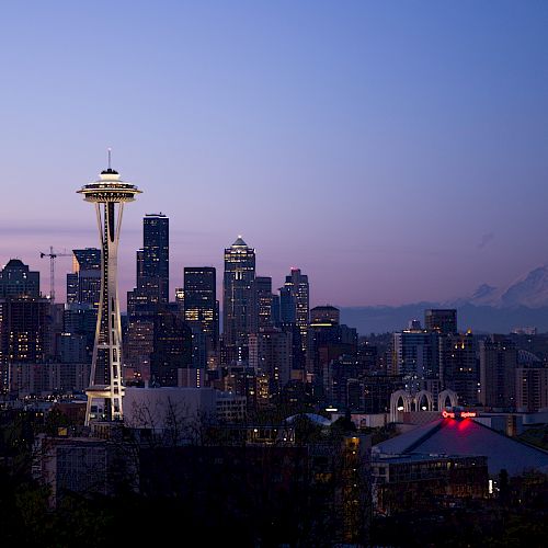 The image shows the Seattle skyline at dusk, featuring the Space Needle and Mount Rainier in the background, with city lights starting to illuminate.