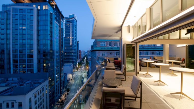 This image shows a modern balcony with seating, overlooking a cityscape with tall buildings, at dusk.