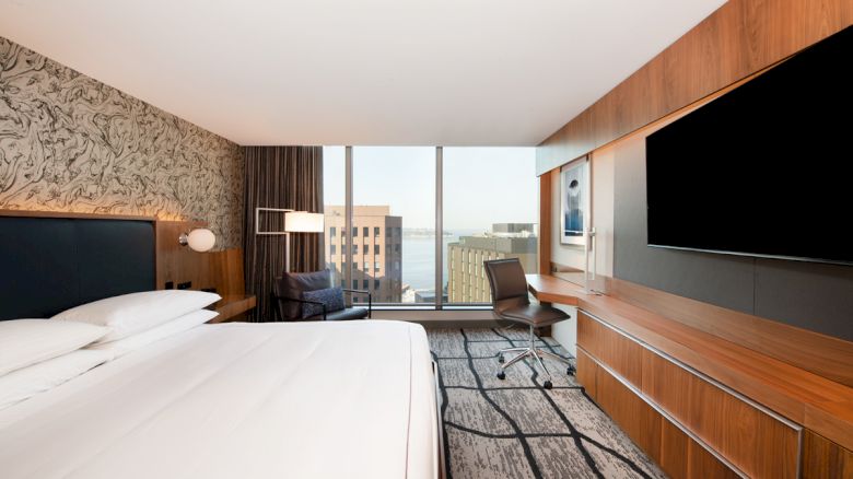 The image shows a modern hotel room with a bed, wall-mounted TV, desk, chair, and large windows offering a city view.