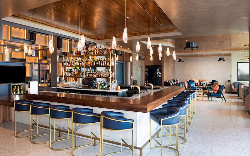 The image shows a modern bar with blue barstools, a well-stocked liquor shelf, and pendant lights above the counter.