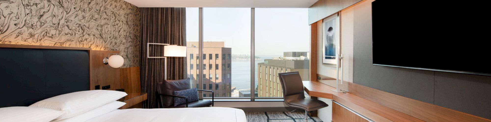 A modern hotel room with a large bed, wall-mounted TV, desk, chair, and a window offering a cityscape view in the background.