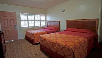 This image shows a simple hotel room with two double beds, a door, windows with blinds, and a patterned carpet.