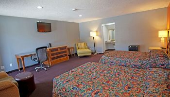 A spacious hotel room with two beds, a desk, a TV, a chair, a small table, and a lamp. An open doorway leads to another room.