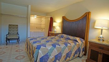 The image shows a hotel room with a bed, nightstand, lamps, an armchair, and an open view of the bathroom area.