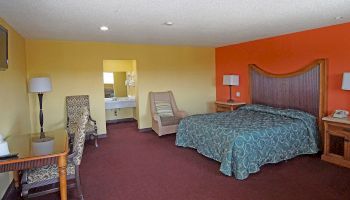 A hotel room featuring a bed, chair, desk with a chair, lamps, a TV, and a sink area visible in the background, with yellow and orange walls.