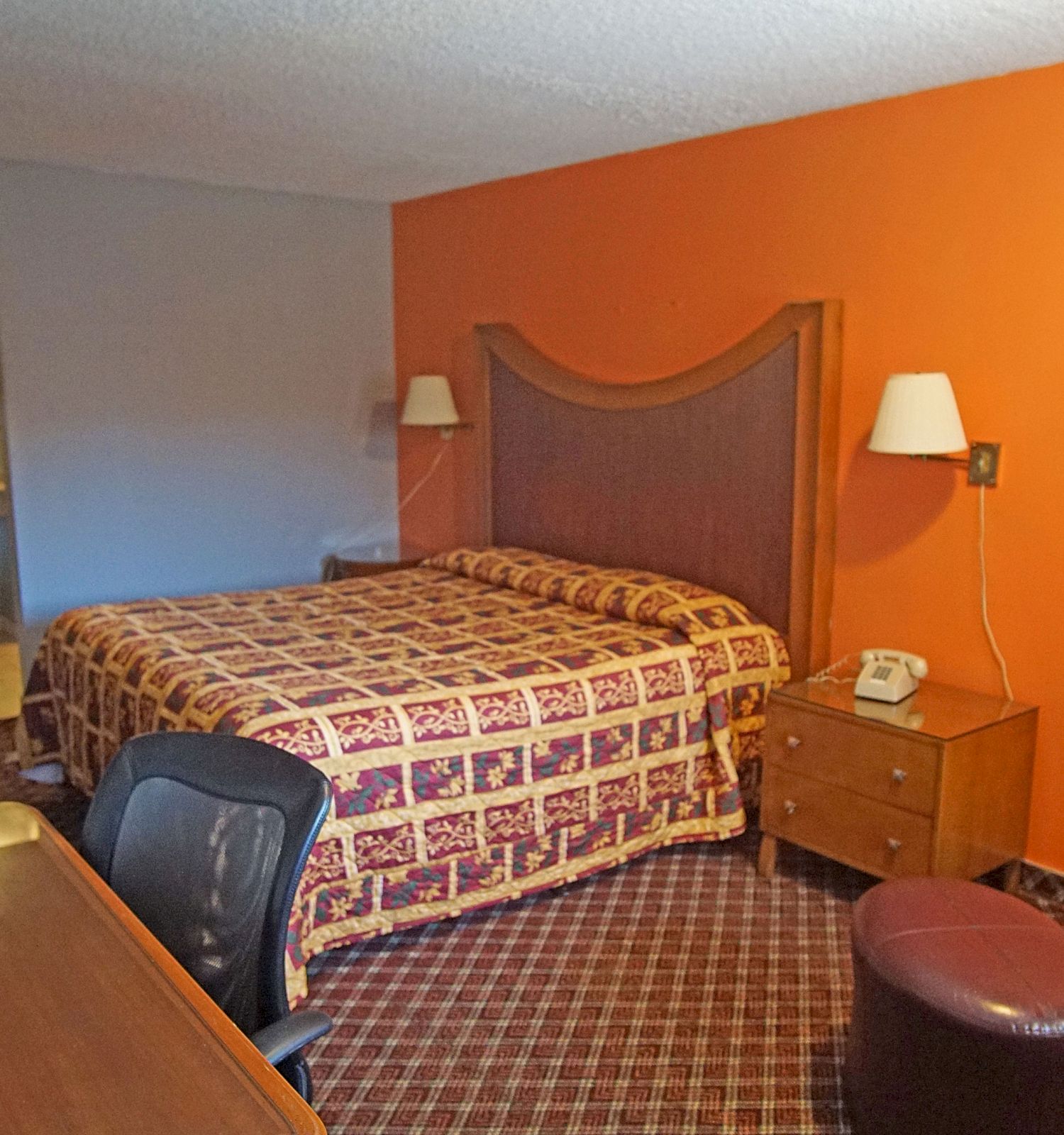 A hotel room with a bed, desk, chair, armchair, ottoman, and bedside table with a phone. The room has an orange accent wall and carpeted floor.