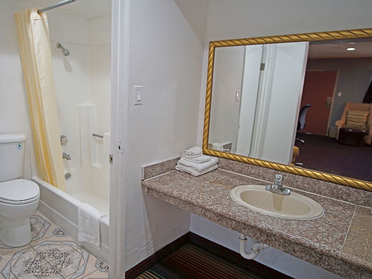 The image shows a bathroom with a toilet, a shower, a large mirror above a sink, and folded towels. Beyond the bathroom is a room with a chair.