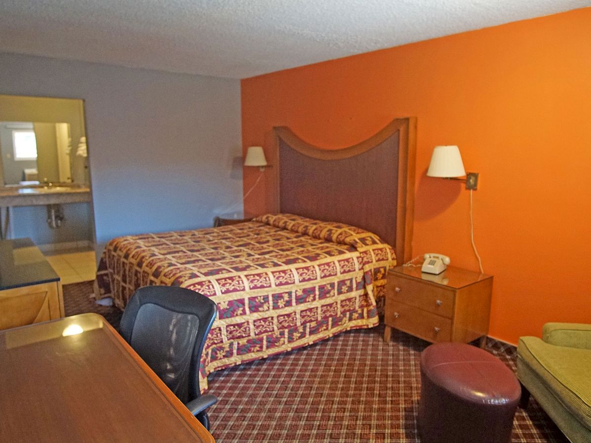 A hotel room with a bed, nightstand, lamps, chair, desk, and a view towards the bathroom in the background, all above a carpeted floor.