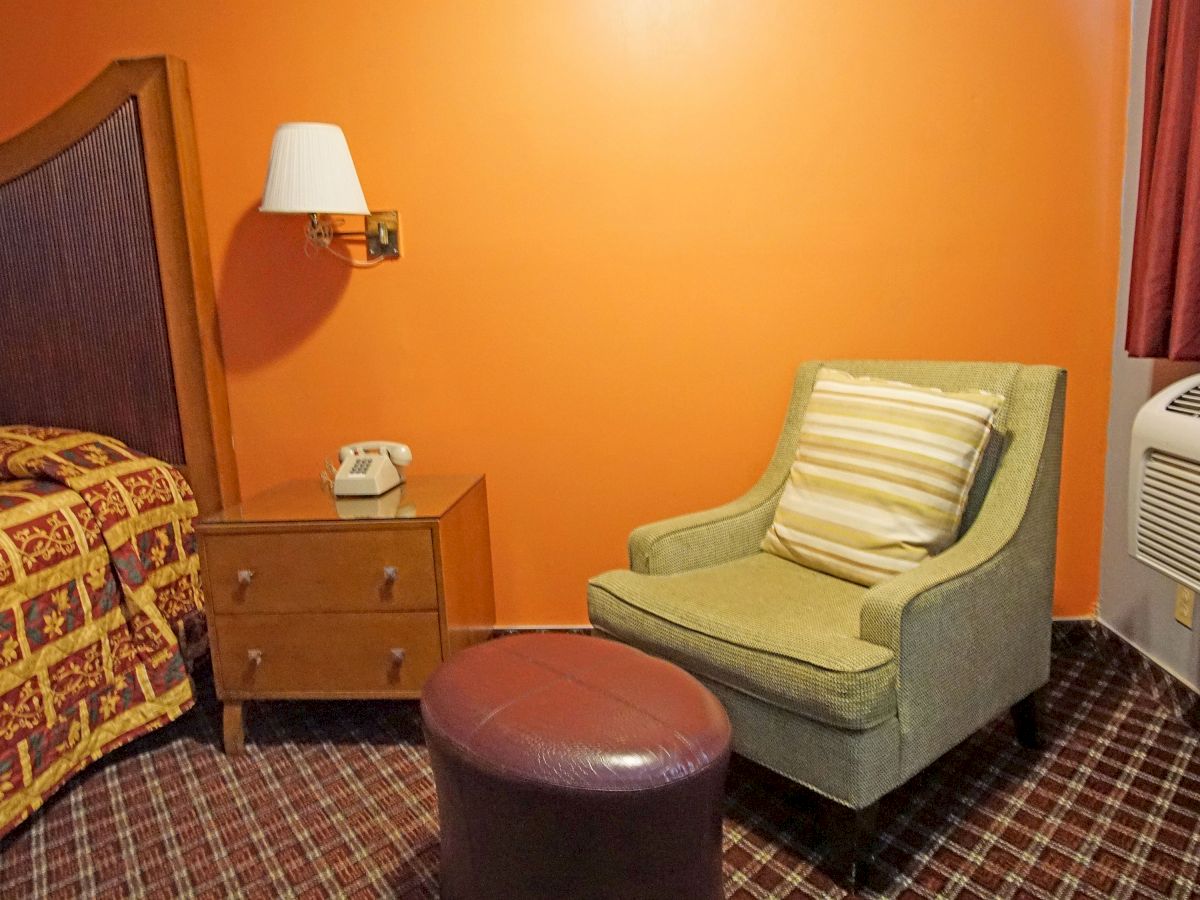 A hotel room showing a bed, nightstand with a phone, upholstered chair with a pillow, round stool, and air conditioning unit.