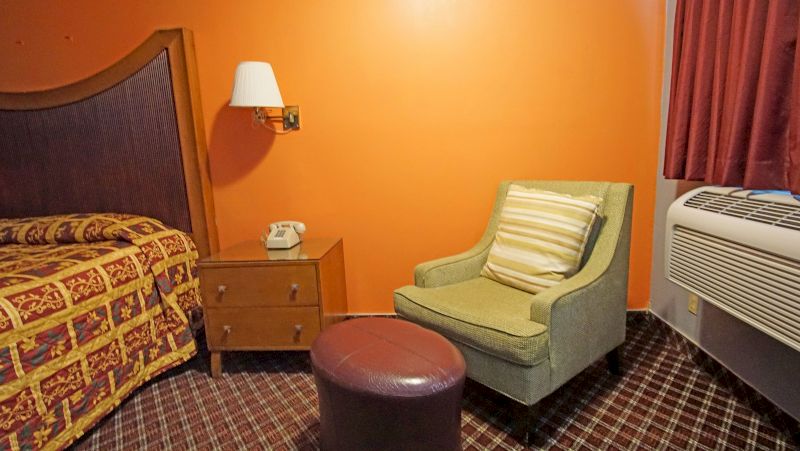 A wide view of a hotel room with a bed, chair, nightstand, and air conditioner.