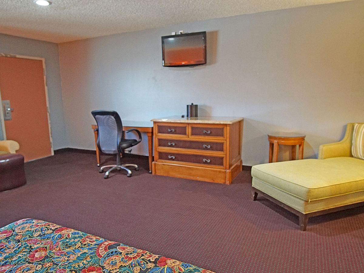 The image shows a hotel room with a desk, chair, couch, armchair, television, and brown carpet. The room has a neutral color scheme.